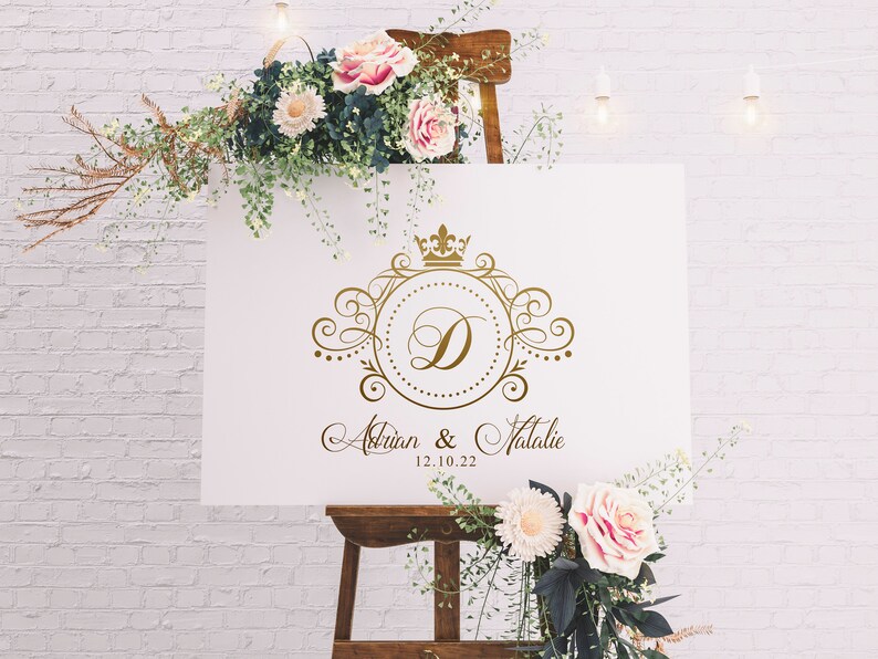 May include: White wedding sign with a gold ornate design featuring a crown and the letter D. The sign reads "Adrian & Natalie 12.10.22".