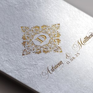 Könnte beinhalten: Eine weiße Karte mit goldener Beschriftung. Die Karte zeigt ein dekoratives Monogramm mit dem Buchstaben "D" in der Mitte. Der Text "Adrienn & Matthew 11.08.25" ist in Kursivschrift geschrieben.