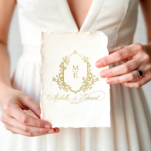 May include: A rectangular off-white card with deckled edges featuring a gold foil-stamped design. The center displays a floral-framed crest with the initials M and E, followed by the names Michelle & Edward and the date 12.14.27 in gold script.