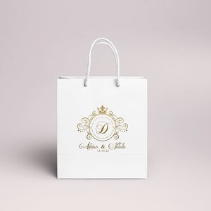 May include: White paper gift bag with a gold crown and the letter D in a circle. The text "Alison & Natalie 12.18.22" is printed below the logo.