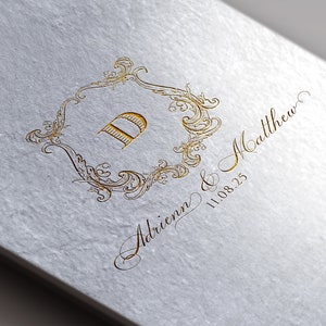Könnte beinhalten: Eine goldfarbene Hochzeitseinladung mit einem dekorativen Rahmen und dem Buchstaben "D" in der Mitte. Der Text "Adrienn & Matthew 11.08.23" ist in Kursivschrift unter dem Rahmen geschrieben.