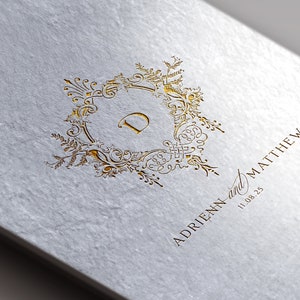 Może przedstawiać: Biała kartka z pozłacanymi napisami. Na kartce znajduje się ozdobny złoty monogram z literą "D" w środku. Pod monogramem widnieje tekst "ADRIEHN and MATTHEW 11.08.25".