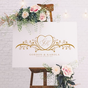Puede incluir: Un cartel blanco con un monograma dorado en forma de corazón con las iniciales "GK" y detalles florales. El texto "Gordon & Karola" y "20th of August 2021" está debajo del monograma.