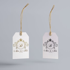 May include: Two white gift tags with gold and black string. Each tag has a decorative design with a crown and the letter D in a circle. The text on the tags reads "Adrian & Natalie 12.08.22".