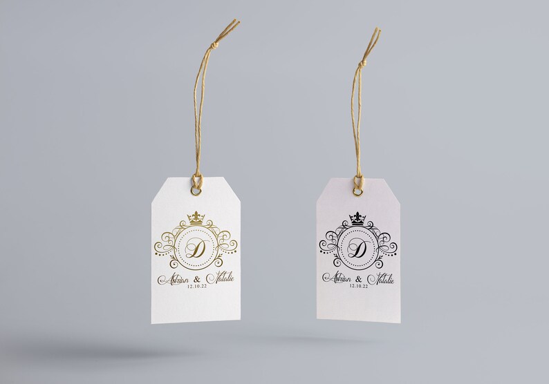 May include: Two white gift tags with gold and black string. Each tag has a decorative design with a crown and the letter D in a circle. The text on the tags reads "Adrian & Natalie 12.08.22".