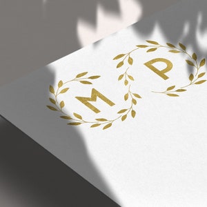 May include: A white card with a gold foil monogram of the letters "M" and "P" surrounded by a wreath of gold leaves.