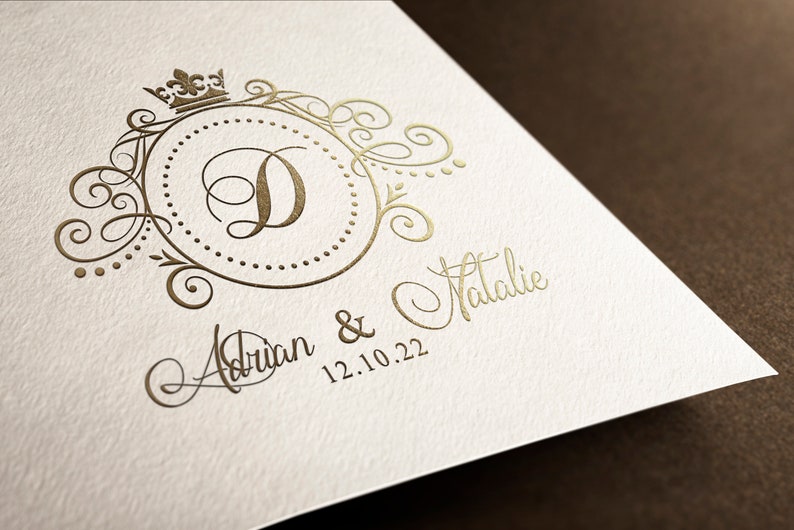 May include: A gold-colored wedding invitation with a decorative monogram featuring the letter "D" and a crown. The text "Adrian & Natalie 12.10.22" is written in cursive below the monogram.