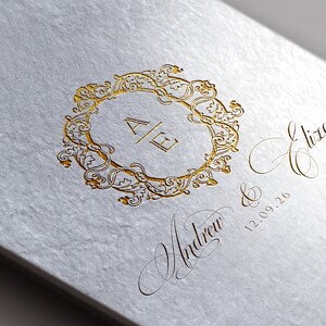 May include: A white card with gold lettering. The card features a decorative gold frame with the initials "A & E" inside. Below the frame, the text reads "Andrew & Eliza 12.09.26".