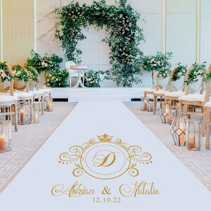 May include: A white aisle runner with a gold monogram featuring the letter "D" and the names "Adrian & Natalie" with the date "12.10.22". The runner is decorated with a gold crown and ornate scrollwork. The runner is placed in a wedding ceremony setting with white chairs and floral arrangements.