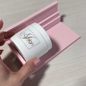 Puede incluir: Un recipiente cilíndrico blanco con una etiqueta que dice "Design Studio" se coloca sobre una plataforma rosa con bordes de regla. La plataforma tiene una base plana y dos secciones elevadas con marcas de medición. Los artículos están sobre una superficie clara.