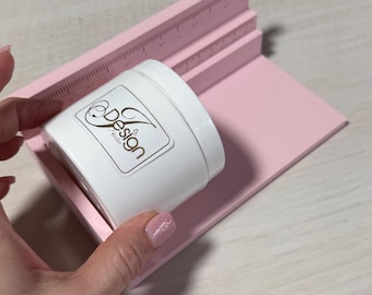 May include: A white cylindrical jar with a label reading Z Design Studio, positioned against a light pink plastic alignment tool featuring a ruler with imperial markings along the top edge to assist with precise placement.
