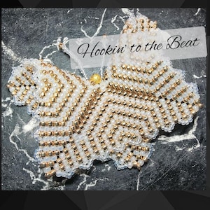 May include: A white and gold beaded butterfly with a gold bead in the center. The butterfly is on a gray and white marble background. The text "Hookin' to the Beat" is above the butterfly.