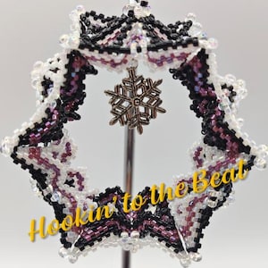 May include: A beaded ornament in a starburst shape, featuring white, black, and purple beads. A silver snowflake charm hangs in the center. The text "Hookin' to the Beat" is written in yellow across the bottom.