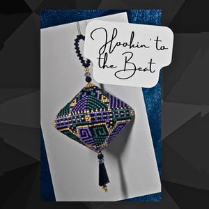 Puede incluir: Un adorno de cuentas de color morado, verde y dorado con un borla negra colgando. El adorno tiene forma de diamante y presenta un patrón geométrico. El adorno está colgado de una cuerda negra y está etiquetado como "Hookin' to the Beat".