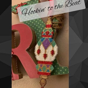 May include: A beaded ornament in the shape of a Christmas tree with red, green, white, gold, and purple beads. The ornament is hanging from a brown rope and the text "Hookin' to the Beat" is visible in the background.