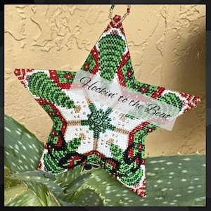 May include: A beaded star ornament with a festive design. The star features a white center with a snowflake pattern, surrounded by green and red accents. The text "Hookin' to the Beat" is displayed across the front. A beaded loop is attached for hanging.