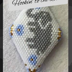Op de afbeelding: Een ruitvormig ornament van kralen met de tekst Hookin' to the Beat. Het ontwerp is gemaakt met witte, grijze, lichtblauwe en metallic gouden kralen in een geometrisch patroon met kleine bloemachtige accenten aan de zijkanten.