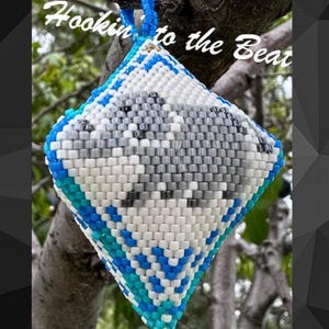 Op de afbeelding: Een witte, grijze, blauwe en turquoise kralenversiering in de vorm van een ruit met de tekst "Hookin' to the Beat" hangend aan een tak.