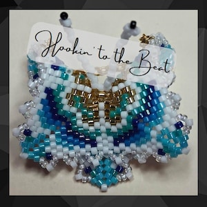 Op de afbeelding: Een kralen vlinder met een wit, blauw en gouden kleurenschema. De vlinder is gemaakt van kleine kralen en heeft een delicaat, ingewikkeld ontwerp. De tekst "Hookin' to the Beat" is in wit op een zwarte achtergrond geschreven.