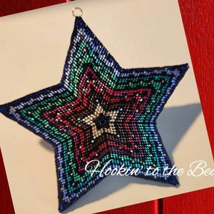May include: A multi-colored beaded star ornament with a blue, green, red, and black color scheme. The star is made of small beads and has a layered design. The ornament is hanging from a gold loop. The text "Hookin' to the Beat" is visible in the bottom right corner of the image.
