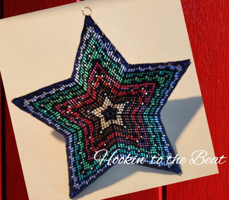 May include: A multi-colored beaded star ornament with a blue, green, red, and black color scheme. The star is made of small beads and has a layered design. The ornament is hanging from a gold loop. The text "Hookin' to the Beat" is visible in the bottom right corner of the image.