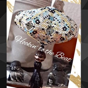 May include: A crocheted, multi-coloured, cloud-shaped ornament with a black tassel and a brown bead. The ornament is hanging from a white string and the text "Hookin' to the Beat" is written below it.