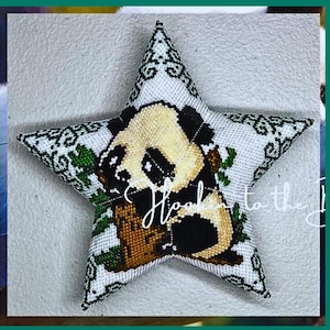 May include: A white star-shaped pillow with a beaded panda bear design. The panda is holding a green bamboo shoot in its mouth. The pillow has a white background with green and white beaded designs around the edges.
