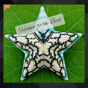 Pode incluir: Um ornamento de contas em forma de estrela branca, verde e azul com o texto "Hookin' to the Beat" sobre um fundo de folha verde.