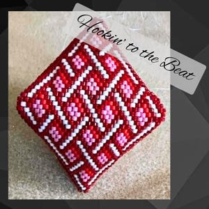 May include: A square, beaded item with a red background and a geometric pattern of white and pink diamonds. The text "Hookin' to the Beat" is in the upper right corner.