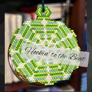 May include: A green, white, and blue beaded ornament with a snowflake design. The ornament is hanging from a silver hook and has the text "Hookin' to the Beat" on it.