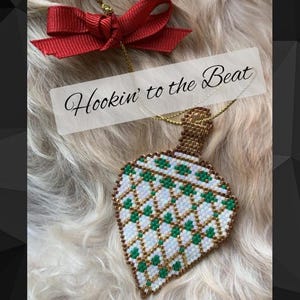 May include: A white and green beaded ornament shaped like a teardrop with gold accents. The ornament is hanging from a gold chain and a red bow. The text "Hookin' to the Beat" is written in white on a black background.