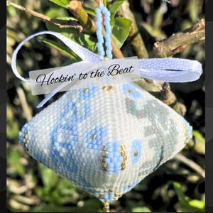 Op de afbeelding: Een met kralen versierde diamantvormige ornament met een witte basis en blauwe en grijze bloempatronen. Het heeft een wit lint en de tekst "Hookin' to the Beat". Gouden kralen accentueren het ontwerp en het hangt aan een blauw kralensnoer.
