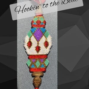 May include: A colorful beaded ornament with a geometric design. The ornament is red, white, gold, blue, green, and purple. The ornament is hanging from a red ribbon and has the text "Hookin' to the Beat" above it.