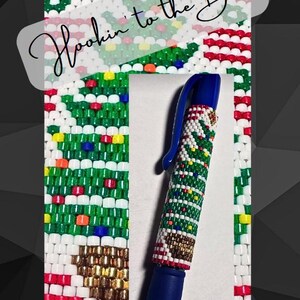 Può includere: Una penna blu con un design a perline di un albero di Natale con perline rosse, verdi, bianche, dorate e gialle. Il testo "Hookin' to the Beat" è visibile sullo sfondo.