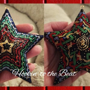 May include: A beaded star ornament with a multi-colored geometric pattern in shades of red, green, gold, and blue. The star is held in a hand and has a gold loop at the top for hanging.