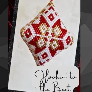 May include: A red, white, and gold beaded ornament with a geometric pattern. The ornament is shaped like a diamond and has a loop at the top for hanging. The text "Hookin' to the Beat" is written below the ornament.