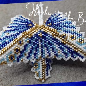 Könnte beinhalten: Ein blauer, weißer und goldener Perlen-Schmetterlings-Anhänger. Der Schmetterling ist aus kleinen, quadratischen Perlen gefertigt und hat ein detailliertes Design.