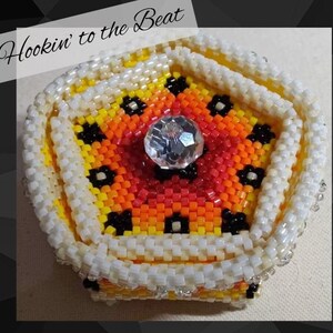 May include: A beaded, star-shaped box with a clear, faceted gem in the center. The box features a gradient of colors, including yellow, orange, and red, with black floral accents. The border is white, and the text "Hookin' to the Beat" is in the upper left corner.