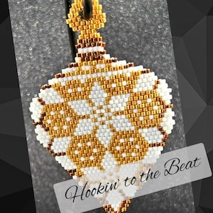 Può includere: Un ornamento a forma di lacrima con un designo a stella, realizzato con perline bianche e dorate. L'ornamento ha un anello in cima per appenderlo e il testo "Hookin' to the Beat" è scritto sotto l'ornamento.