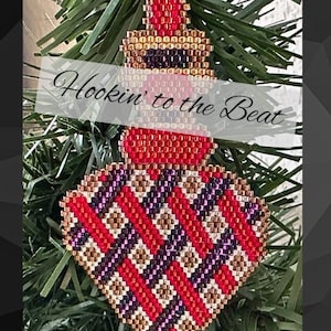 May include: A beaded Christmas ornament in the shape of a heart with a geometric pattern of red, gold, and purple beads. The ornament is hanging on a green Christmas tree branch. The text "Hookin' to the Beat" is visible in the image.