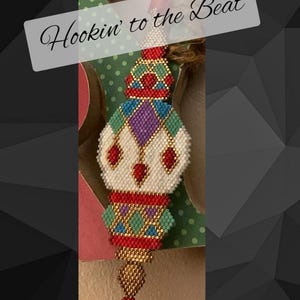 May include: A colorful beaded ornament with a geometric design. The ornament is white, red, green, gold, and purple. The ornament has a gold tassel at the bottom. The text "Hookin' to the Beat" is written on a white background.