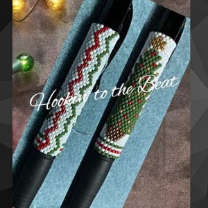 May include: Two beaded pens with Christmas themes. One pen has a red and white chevron pattern, the other has a green Christmas tree with red baubles. The pens are black and have the text "Hookin to the Beat" on the image.