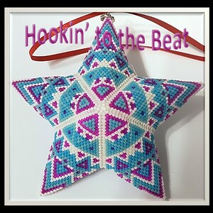 May include: A beaded star ornament in shades of blue, pink, and white. The star is 3D and has a geometric pattern. The ornament is hanging from a red ribbon with the text "Hookin' to the Beat" on it.