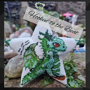 Könnte beinhalten: Ein weißer, sternförmiger Schmuck mit einem Perlendesign, das einen grünen Drachen zeigt. Der Drache hat rote Augen und ist mit verschiedenen Grüntönen detailliert. Der Schmuck trägt den Text "Hookin' to the Beat".