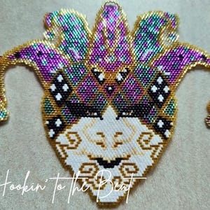 May include: A beaded jester mask in shades of purple, teal, gold, black, and white. The mask features a diamond pattern and decorative swirls. The text "Hookin' to the Beat" is written in cursive at the bottom.
