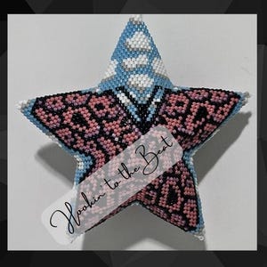 Pode incluir: Uma estrela de contas rosa e preta com um centro de contas azuis e brancas. A estrela tem uma etiqueta branca com o texto "Hookin' to the Beat" escrito em preto.