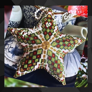 Może przedstawiać: Gwiazdkowa ozdoba z koralików z geometrycznym wzorem w kolorze zielonym, czerwonym i złotym. Ozdoba ma białe tło, a na niej napis "Hoodie to the Bed".