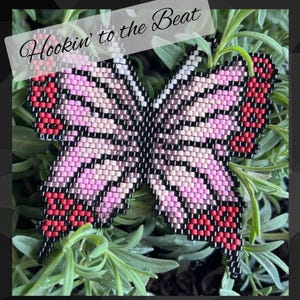 May include: A pink and black beaded butterfly with red accents. The butterfly is sitting on a bed of green foliage. The text "Hookin' to the Beat" is written in white on a black background.
