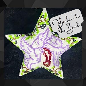 May include: A white star-shaped ornament with a purple and red design. The ornament is decorated with green and brown beads. The text "Hookin' to the Beat" is written on a white tag.