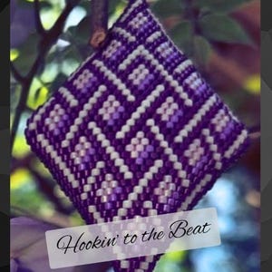 May include: A handmade, diamond-shaped ornament with a beaded design in shades of purple and white. The ornament is suspended from a small branch against a blurred background of green leaves. The text "Hookin' to the Beat" is displayed on the bottom.